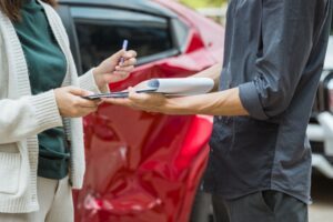 Two people looking over paperwork with a damaged car in the background.
